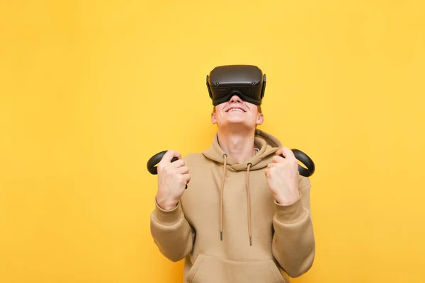 Le gars heureux dans le casque VR se réjouit de gagner sur un fond jaune, porte des vêtements décontractés et tient les contrôleurs de jeu dans sa main.Jeune homme jouant jeu VR dans le casque de réalité virtuelle, satisfait de regarder vers le haut . — Photo
