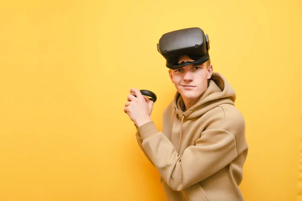 Joueur VR drôle avec casque sur la tête et contrôleur dans ses mains se tient sur un fond jaune, regarde dans la caméra et sourit. Portrait VR de gamer adolescent isolé. Espace de copie — Photo