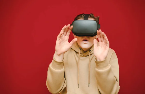 Portrait d'un gars choqué dans un casque VR sur un fond rouge, couvre son visage avec sa main et regarde en caméra avec un visage surpris, portant des vêtements décontractés. adolescent émotionnel utilise des lunettes VR — Photo