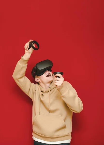 Le gars en tenue décontractée et un casque VR joue à des jeux de réalité virtuelle sur fond rouge, regarde l'espace flic avec un visage émotionnel et se réjouit. Adolescent joyeux jouer à des jeux VR avec contrôleur dans son — Photo