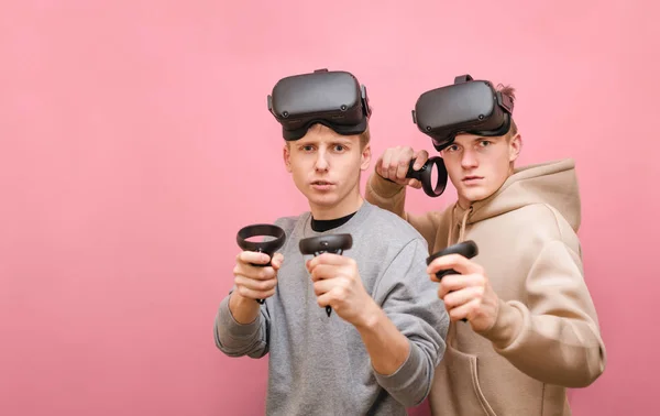 Portrét dvou legračních přátel v helmě na růžovém pozadí hledících do kamery s vážnou tváří. Dva kluci v neformálním oblečení hrát Vr hry s regulátory v jejich rukou — Stock fotografie
