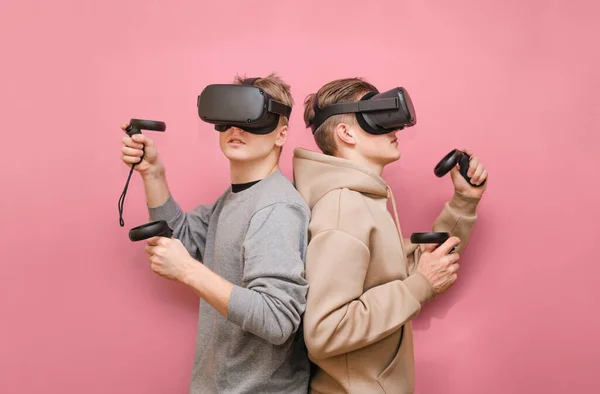 Two young men in VR helmets on their heads stand side by side on a pink background with controllers in their hands. Team of guys playing VR games together, isolated. VR gaming with friends.