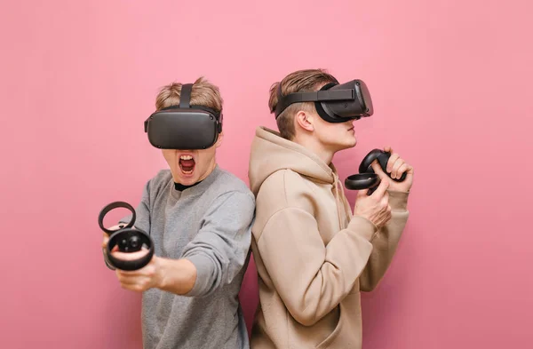 Casal de amigos engraçados usando capacetes VR e controladores nas mãos, de pé sobre um fundo rosa e jogando jogos de realidade virtual. Dois jovens em roupas casuais jogando jogos em VR — Fotografia de Stock