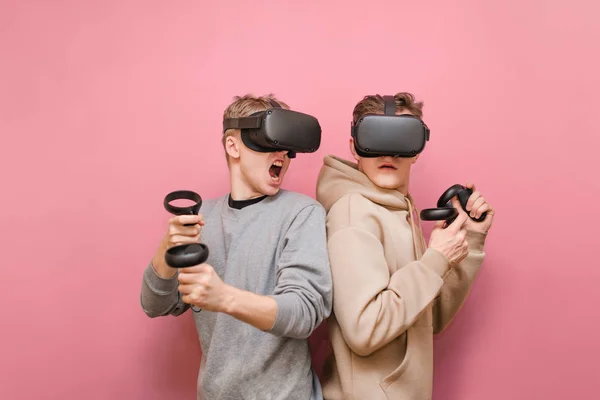 Deux jeunes hommes émotionnels dans les casques VR concourent dans un jeu de réalité virtuelle. Deux joueurs dans les casques de réalité virtuelle jouent à des jeux VR, tenir les contrôleurs. VR jeux, loisirs avec des amis — Photo