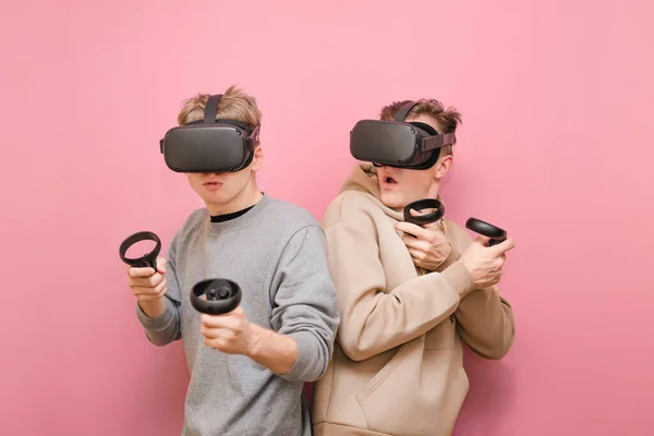 Deux drôles de joueurs en casques VR sur fond rose, jouant ensemble à des jeux de réalité virtuelle et réagissant émotionnellement. Deux jeunes hommes isolés sur fond rose jouant à des jeux VR sur contrôleurs — Photo