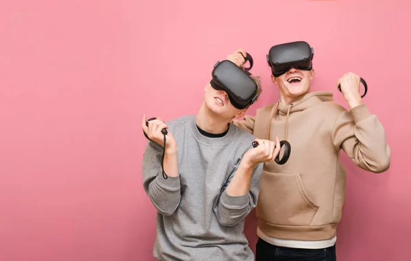 Dva veselí přátelé stojící na růžovém pozadí s helmami Vr na hlavách a ovladači v rukou a hraní videoher ve virtuální realitě. Dva kluci hrají Vr hry v helmách virtuální reality — Stock fotografie