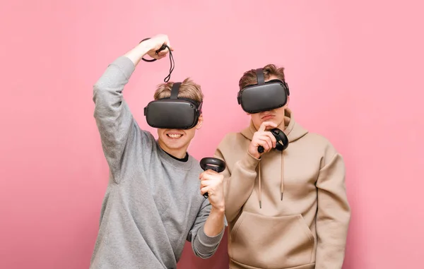 Deux amis émotionnels passent du temps à jouer à des jeux VR, portant des casques de réalité virtuelle et des contrôleurs dans leurs mains et en compétition. Deux gars dans des casques VR isolés sur fond rose . — Photo