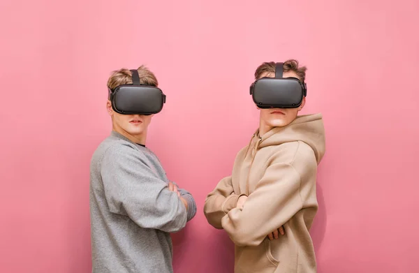 Portrait de deux jeunes hommes en vêtements décontractés debout sur un fond rose avec des casques VR sur la tête, regardant dans la caméra avec un visage sérieux. Deux amis utilisent des casques de réalité virtuelle, isolés — Photo