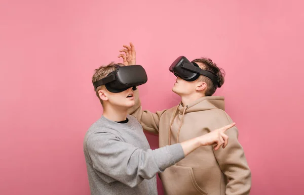 Deux gars surpris dans des lunettes VR se tiennent sur un fond rose regardant loin et pointant leur doigt. Deux autres regardent dans un casque de réalité virtuelle, ils sont choqués. Espace de copie — Photo