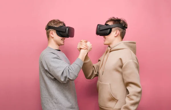 Deux jeunes hommes heureux portant un casque VR se regardent et se serrent la main avec un sourire sur le visage. Deux joyeuses équipes de joueurs dans les casques VR rivalisent sur fond rose. Jeux VR . — Photo