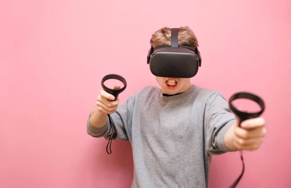 Portrait de jeune homme expressif en casque VR et avec des contrôleurs dans les mains sur fond rose regardant dans la caméra avec un visage fou. Homme émotionnel jouant à des jeux VR — Photo