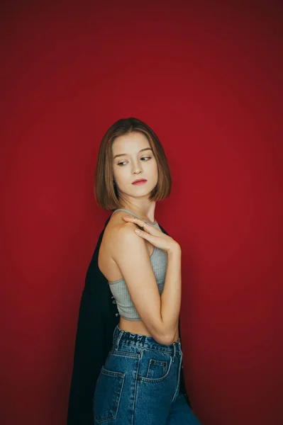 Modelo de menina sexy em jersey e jeans fica no fundo vermelho e olha para o lado, posando para a câmera. Isolado. Retrato vertical de menina sexy no fundo da parede vermelha . — Fotografia de Stock