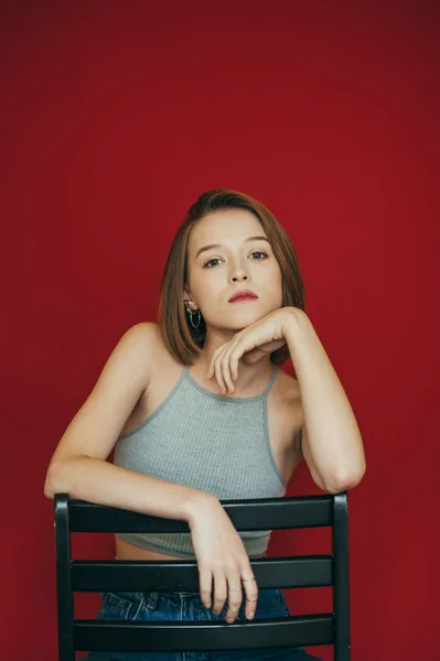 Modelo atraente em uma camiseta branca senta-se em uma cadeira contra um fundo vermelho e posa para a câmera com um rosto sério. Senhora bonita em roupas casuais na cadeira isolada no fundo vermelho.Vertical — Fotografia de Stock