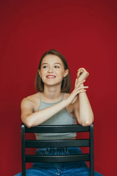 Positief meisje in een grijs shirt zit op een stoel op een rode achtergrond, kijkt weg met een glimlach op haar gezicht. Gelukkig model in casual kleding poseren op stoel op rode achtergrond. Verticale foto — Stockfoto