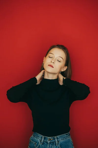 Attractive girl in a dark sweater and jeans stands on a red background with her eyes closed, poses for the camera and enjoys. Portrait of cute girl model with closed eyes on isolated on red background — 스톡 사진