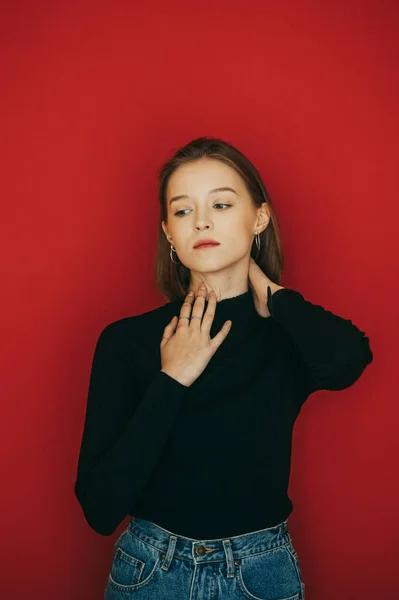 Lindo modelo en ropa casual oscura se levanta sobre un fondo rojo, posa para la cámara y mira hacia otro lado. Elegante dama atractiva aislada sobre fondo rojo. Vertical . —  Fotos de Stock