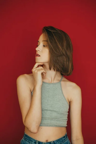 Aantrekkelijk model in stijlvolle zomerkleding poserend op rode muurachtergrond. Sexy meisje in casual kleding, draagt lichte t-shirt en jeans, geïsoleerd op rode achtergrond, kijkt weg met ernstige gezicht. — Stockfoto