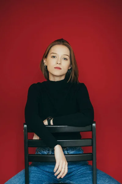 Chica encantadora en jeans y un suéter negro se sienta en una silla sobre un fondo rojo, mira a la cámara con una cara seria. Modelo atractivo posando en una silla sobre un fondo rojo en el estudio . — Foto de Stock