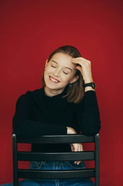 La modelo de la muchacha feliz se sienta en una silla con sus ojos cerrados en el estudio sobre un fondo rojo y sonríe sinceramente.La señora alegre en suéter negro se sienta en una silla contra un fondo de pared rojo. Foto vertical —  Fotos de Stock