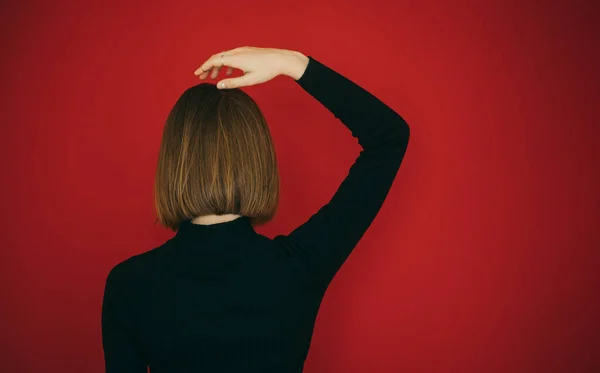 Girl with bob hairstyle stands with her back against a red background and straightens hair. Woman with short hair isolated on red, bob hairstyle. Copy space. Background. — Stock Photo, Image