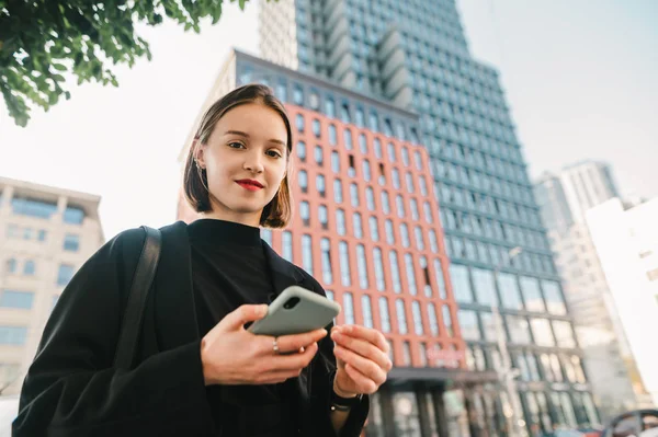 Πορτρέτο ενός όμορφου κοριτσιού με ένα smartphone στα χέρια της στέκεται στο δρόμο μιας μητρόπολης στο παρασκήνιο ενός κτιρίου ουρανοξύστη, κοιτάζει στην κάμερα και χαμογελά. Αντιγραφή χώρου — Φωτογραφία Αρχείου