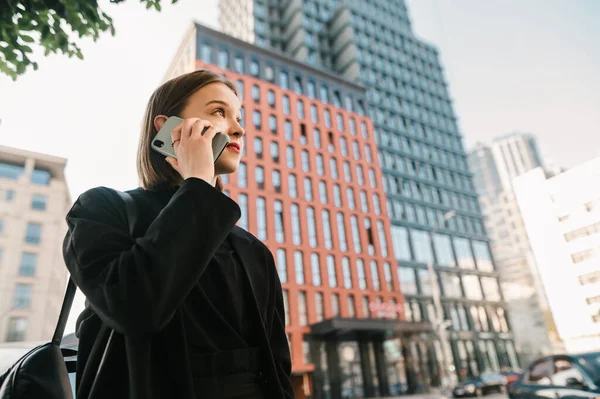 Chica atractiva en una chaqueta y rbkzak se encuentra en la calle de una gran ciudad y se comunica por teléfono. Joven mujer de negocios de moda llamando sobre un fondo de rascacielos. Copiar espacio — Foto de Stock