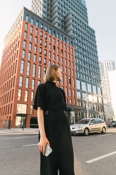 Foto vertical de uma senhora elegante em roupas escuras fica com um smartphone contra o pano de fundo da arquitetura moderna e uma rua metropolitana, olhando para o lado. Estilo de vida retrato de menina na moda — Fotografia de Stock