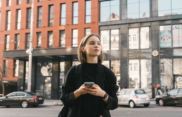 Φωτογραφία δρόμου ενός κομψού κοριτσιού με σακάκι που στέκεται στο δρόμο της πόλης με ένα smartphone στο χέρι του και κοιτάζει αλλού. Αστικός τρόπος ζωής φωτογραφία της μόδας νεαρή επιχειρηματίας χρησιμοποιώντας το τηλέφωνο. — Φωτογραφία Αρχείου