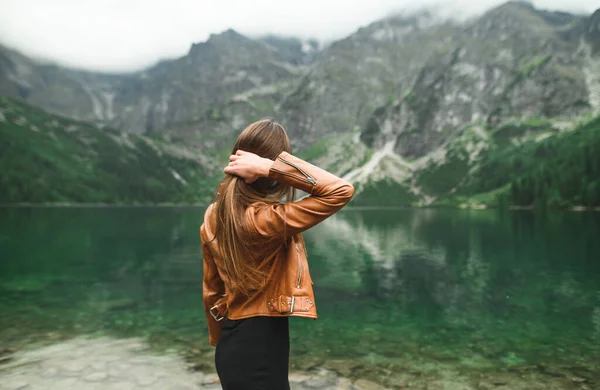 Chica con estilo en ropa casual casual se encuentra en la orilla de un lago de montaña y mira el paisaje irreal. Foto de una chica de atrás en la orilla de un hermoso lago . — Foto de Stock