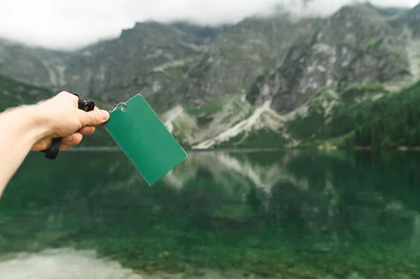 Mão masculina segurando um folheto em branco verde no fundo de um belo lago de montanha espelho. Mapa em branco no fundo do Lago Morskie Oko, Montanhas Tatra. Em branco. Espaço de cópia — Fotografia de Stock