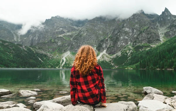 Curly red-haired girl in a red shirt sits on the rocks against the backdrop of a beautiful mountain lake and looks at the landscape.Female tourist sits on the shore of Lake Morskie Oko,Tatra Mountains