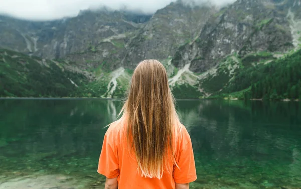 Позаду молодої дівчини-туриста в помаранчевому футболі стоїть на тлі озера в горах і дивиться на нереальний пейзаж. Блондинка на дні озера Морскі Око.. — стокове фото
