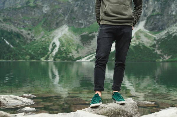 Hiker legs standing on rocks on lake in mountains. A man in sneakers and a green sweater stands on the shores of lake Morskie Oko. Background. Hiker man legs on a hike.