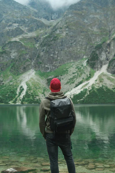 Vertical photo of a man standing on the shores of Lake Morskie Oko in the Tatra Mountains and looking at the beautiful scenery, enjoying, wearing casual clothes and a backpack. Background. — 스톡 사진