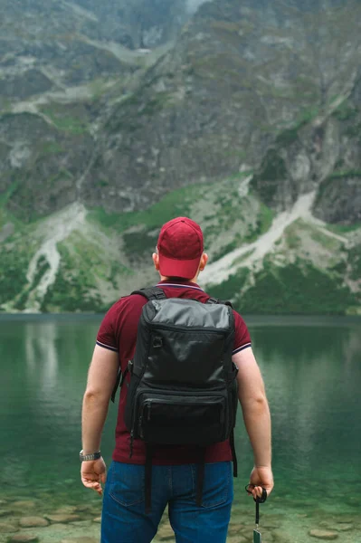 Человек в красной кепке и рюкзаке стоит спиной на озере в горах и смотрит на нереальный пейзаж. Вертикальное фото. Фон. Морские Око. Татранские горы — стоковое фото