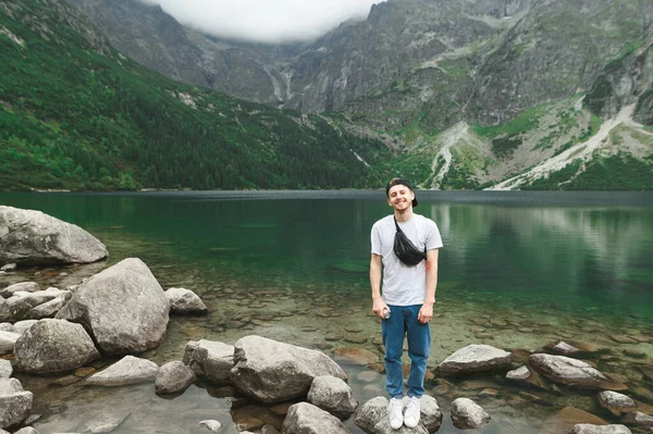 Sonriente joven con gorra y ropa casual se levanta contra el telón de fondo del lago Morskie Oko en las montañas Tatra, mira a la cámara y sonríe. Senderista feliz lago en las montañas — Foto de Stock