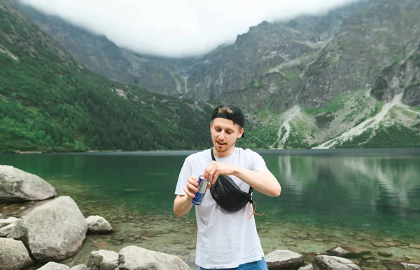 Cara positiva em um boné e t-shirt branca fica nas margens do Lago Morskie Oko e abre uma lata com uma bebida no fundo da paisagem da montanha. Jovem alegre caminhadas em Tatra Mountains — Fotografia de Stock