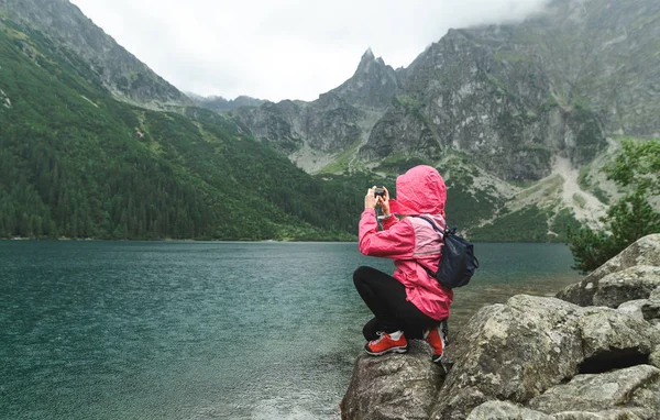 Девушка-туристка сидит на камне на озере в горах под дождем и фотографирует нереальные пейзажи на камеру смартфона, одетая в розовый дождевик красные треккинговые ботинки. Морские Око, Морские глаза — стоковое фото