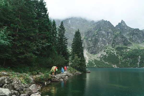 Grupa turystów w płaszczach przeciwdeszczowych spaceruje kamienną ścieżką w pobliżu górskiego jeziora, w tle pięknych skalistych gór. Szlaki krajobrazowe z pięknym widokiem na jezioro Morskie Oko w Tatrach — Zdjęcie stockowe