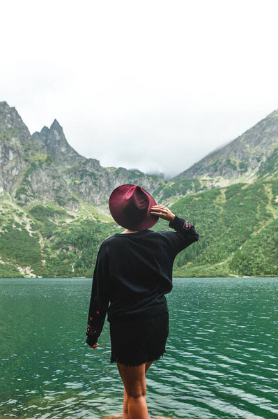 Vertical portrait of a stylish woman in a hat stands with her back against the background of a beautiful mountain lake and cloudy sky. Fashionable woman in a dark dress posing on the shores of Lake