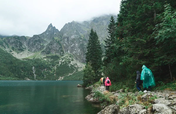 Grupa turystów w płaszczach przeciwdeszczowych spaceruje ścieżką skalną w pobliżu pięknego Jeziora Morskiego Oko z czystą wodą na skalistym tle Tatr. Turyści i wycieczki w deszczu. — Zdjęcie stockowe