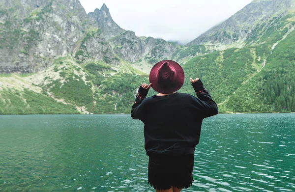 Costas de uma menina elegante em roupas escuras em um fundo de um lago de montanha com água azul e montanhas com nuvens. Contexto. Hipster mulher turística nas margens do Lago Morskie Oko — Fotografia de Stock