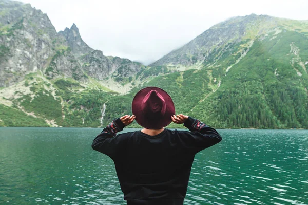 Chica en vestido y sombrero está de pie con su espalda contra el fondo de un gran lago de montaña y posa en la cámara. Hipster retrato de chica en ropa elegante en la naturaleza sobre fondo paisaje irreal — Foto de Stock