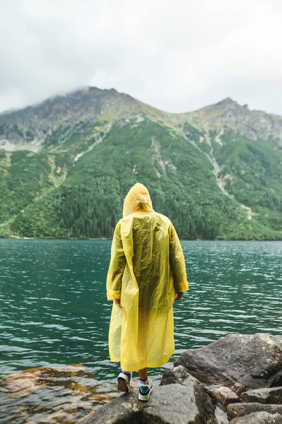 Человек в жёлтом плаще стоит на камне на фоне горного озера Морские Око в Поланде. Пеший туризм возле озера под дождем, стоя спиной на фоне прозрачного озера . — стоковое фото