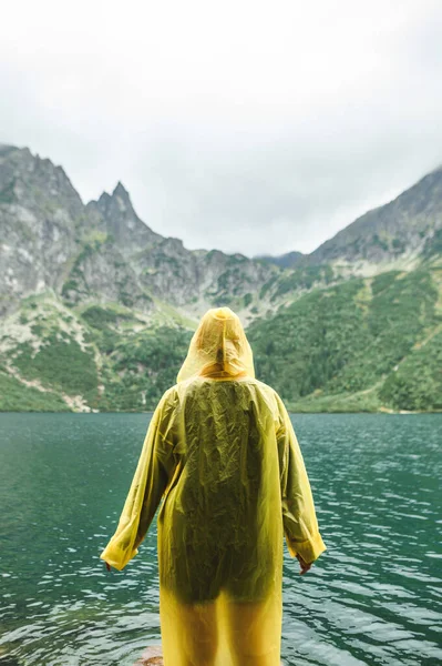 Kontekst. Pionowe zdjęcie. Osoba w żółtym płaszczu przeciwdeszczowym stoi na tle pięknego krajobrazu z Jeziorem Morskim Oko w Tatrach, podziwiając widok. Góry turystyczne — Zdjęcie stockowe