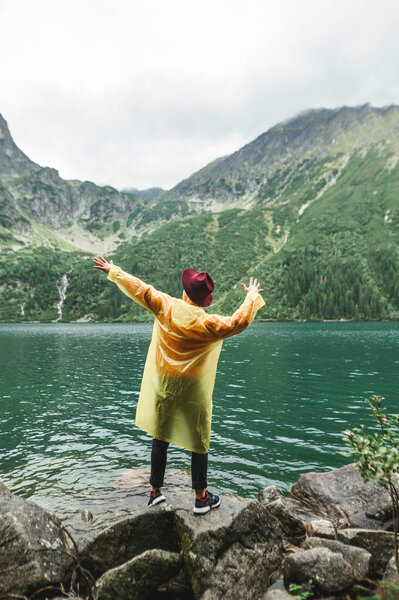 Stylish young man in hat and raincoat stands against blue lake and mountains background and poses at camera. Hipster posing on background of mountain lake Morskie Oko, Tatra Mountains.Vertical photo.