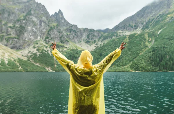 Joyful person in yellow raincoat with raised hands stands against unreal mountain landscape with lake. Person hiking on Lake Morskie Oko in Tatra National Park. Background. Copy space