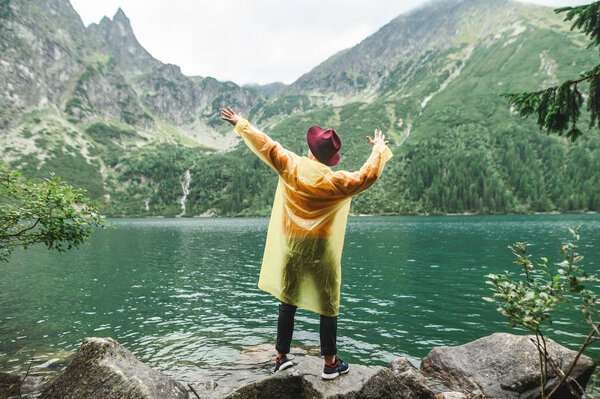 Portrait of a stylish man in a yellow raincoat and hat stands on rocks by the lake overlooking the mountains and shows a Dab. Hipster tourist hiking on a mountain lake, posing for the camera.
