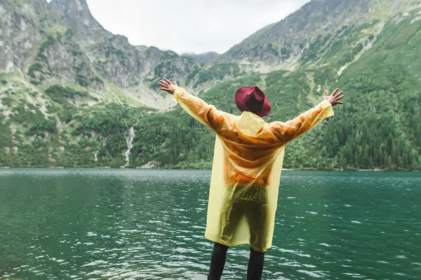 Hombre alegre turista en sombrero y impermeable amarillo se encuentra en la orilla de un lago de montaña con los brazos levantados y disfruta de una vista del paisaje. Antecedentes Caminante excursionista disfrutando del paisaje . — Foto de Stock