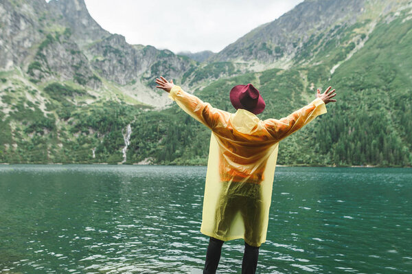 Joyful man tourist in hat and yellow raincoat stands on the shore of a mountain lake with arms raised and enjoys a view of the landscape. Background. Hiker hiking man enjoying the landscape.